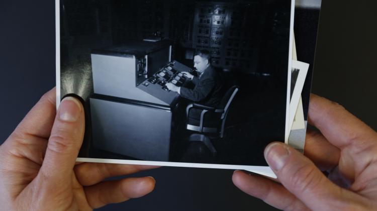 Light-skinned hands hold a black and white photograph of a light-skinned, male-presenting person at a control console in what appears to be the early 20th century.